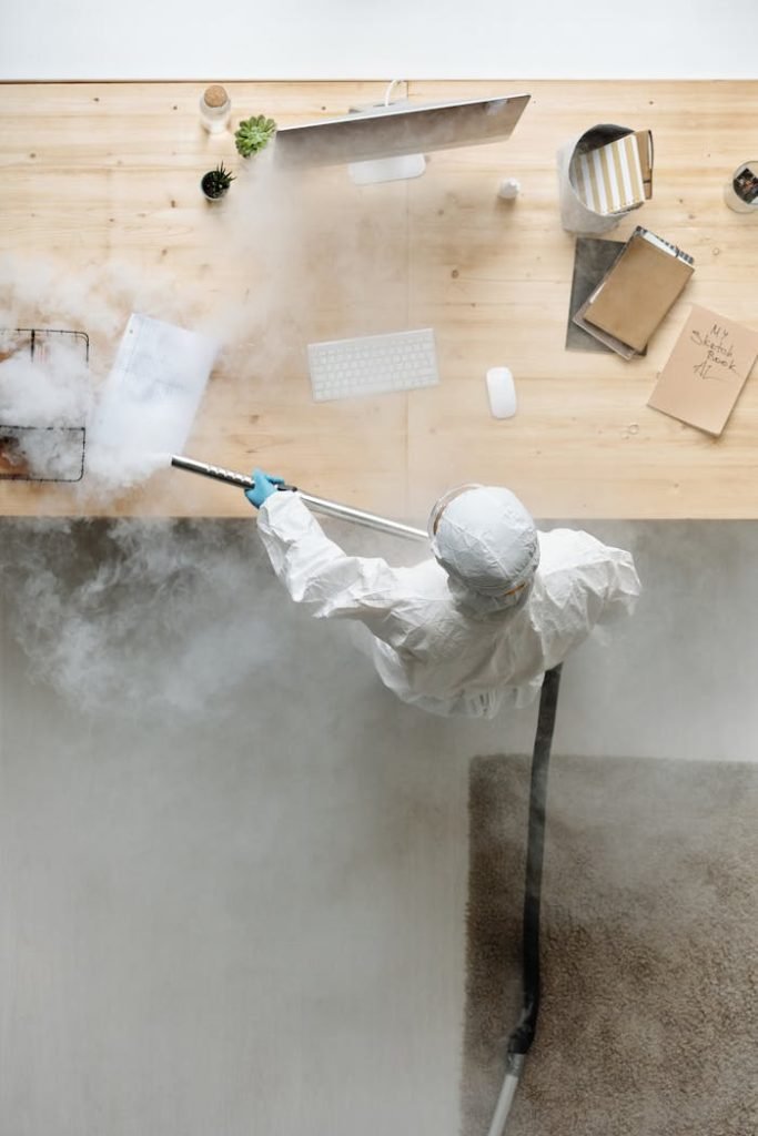 A person in PPE disinfects an office desk with steam, ensuring cleanliness and safety.